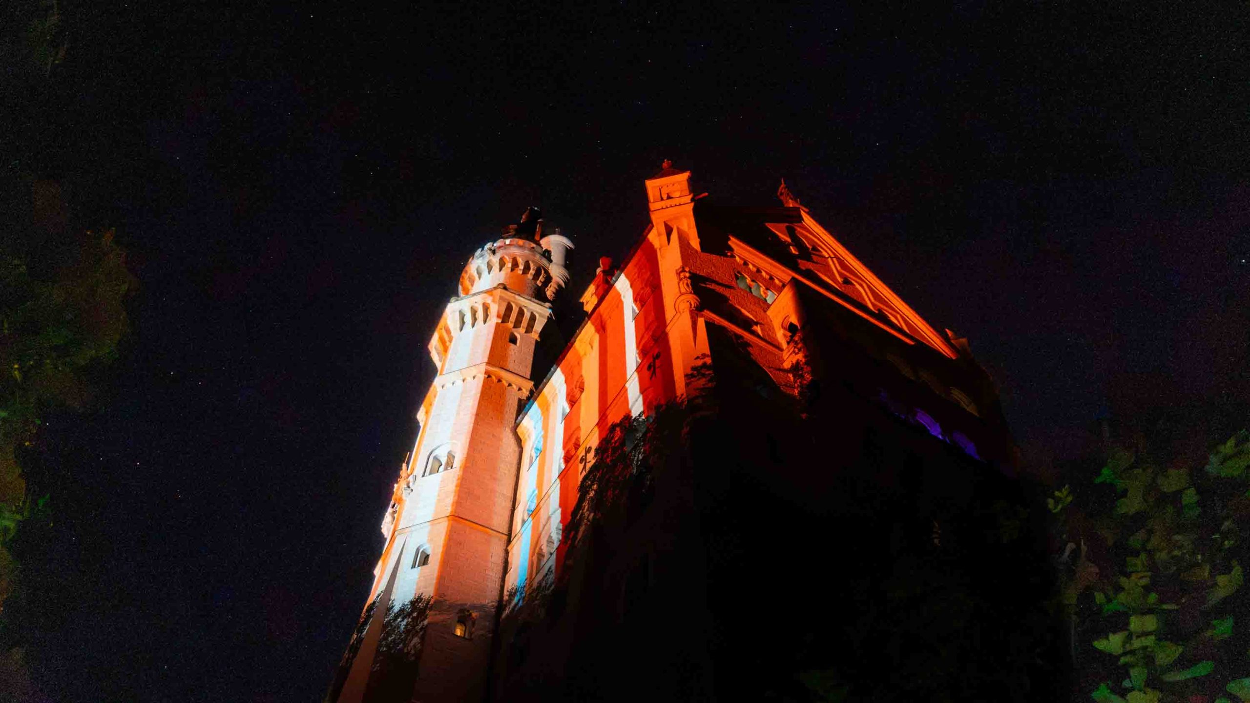 „Neuschwanstein Castle at night, lit in the blue and red climate stripes of Bavaria”