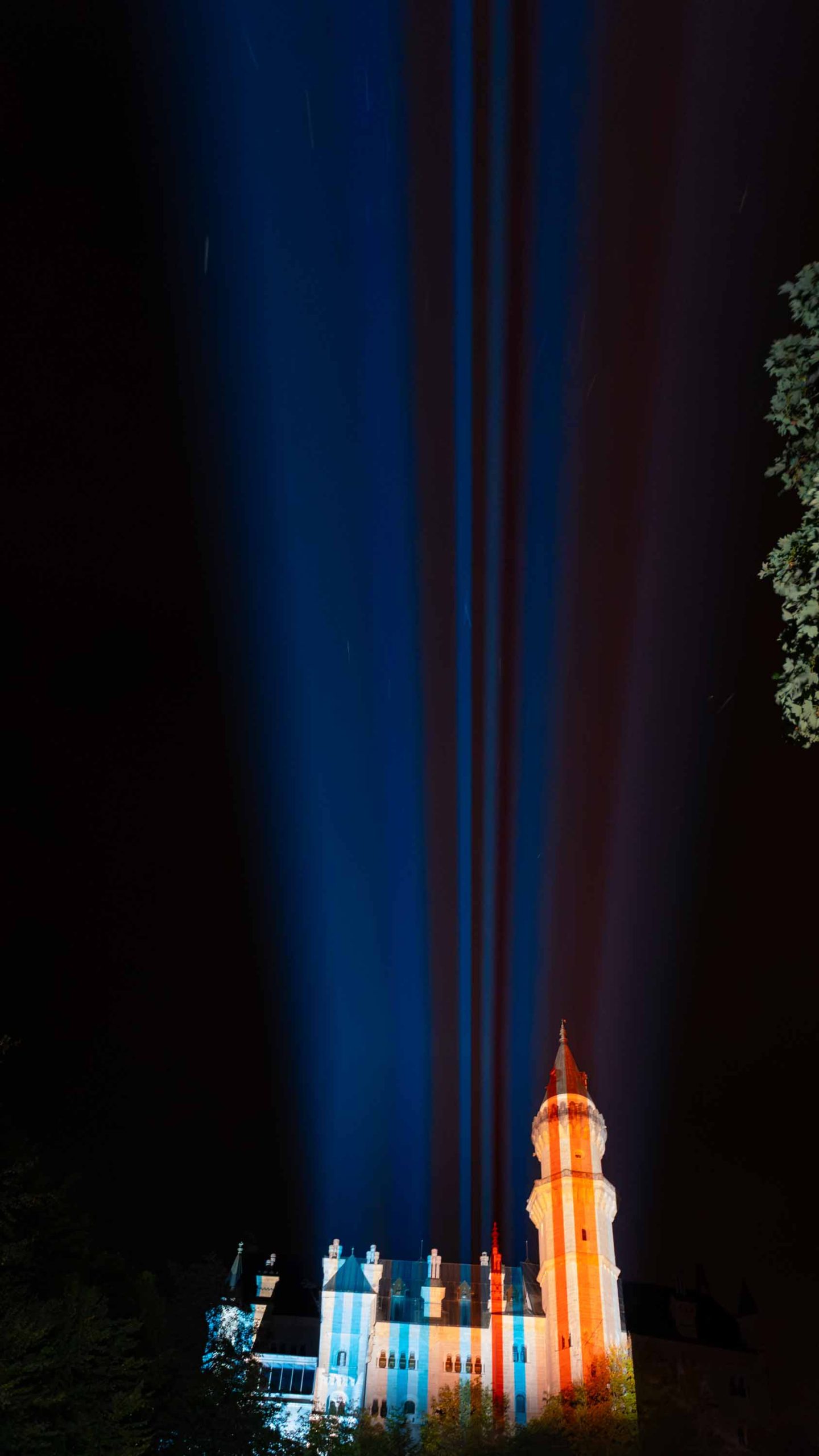 „Neuschwanstein Castle at night, lit in the blue and red climate stripes of Bavaria”